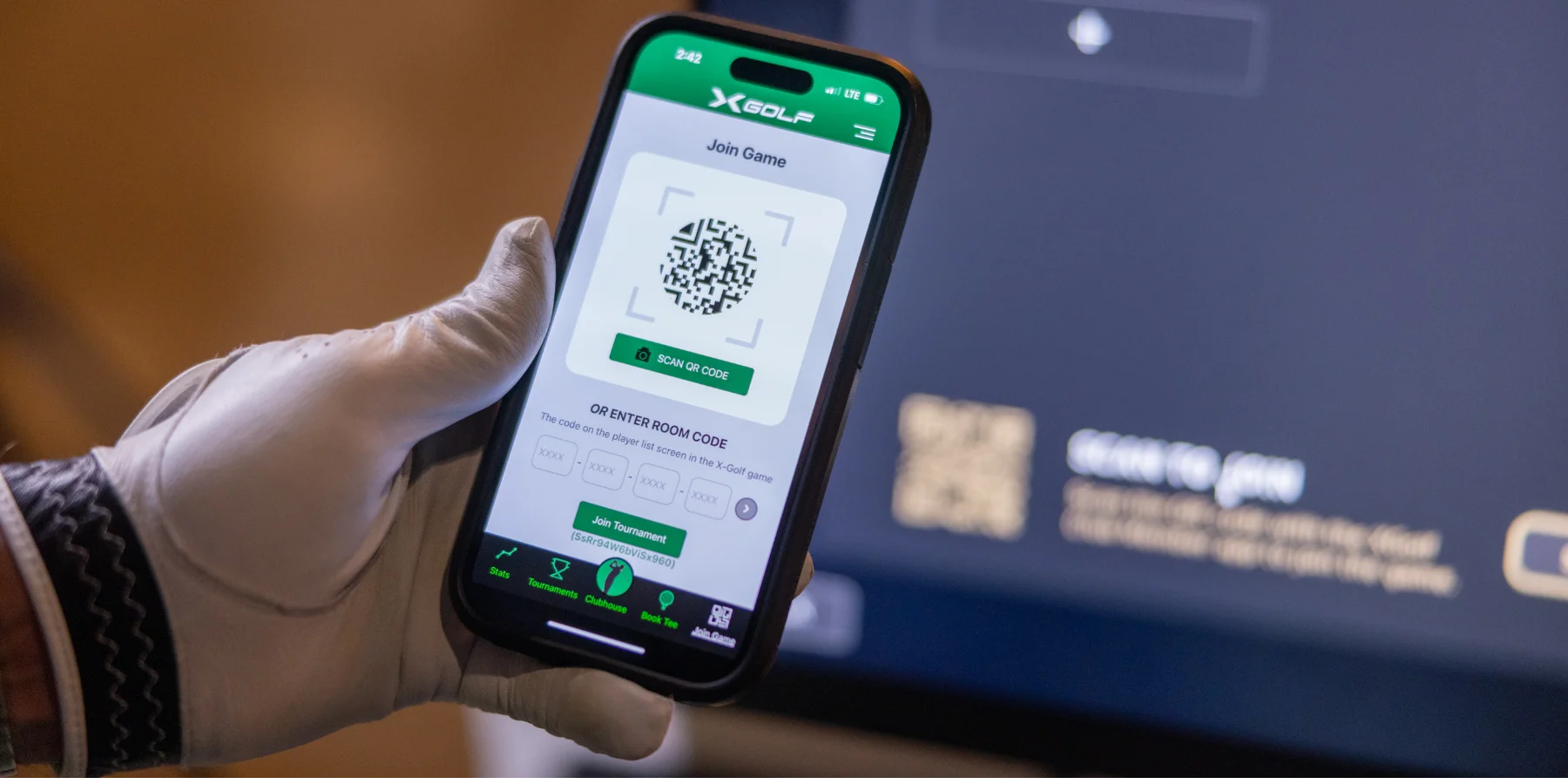 A hand wearing a golfing glove holds a phone that shows the X-Golf app.